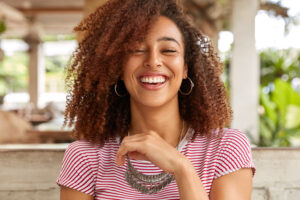 Headshot of glad woman laughs and smiles broadly, expresses positive emotions, has hilarious expression, wears round big earrings and necklace, spends time in cafe. Afro American teenager indoor